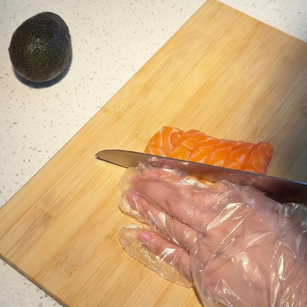 Primo piano di una mano con guanto monouso che usa un coltello da chef per tagliare un filetto di salmone venato a cubetti su un tagliere di bambù.
