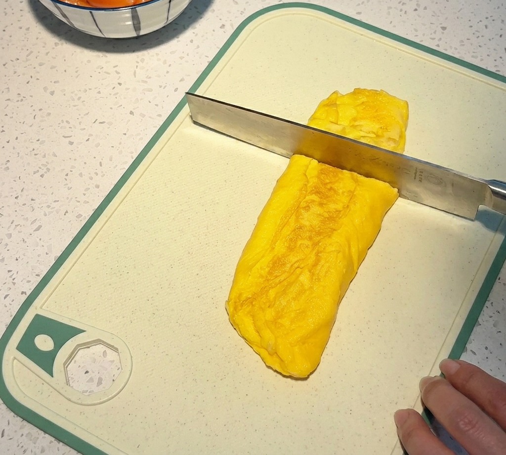 Un coltello taglia una spessa frittata arrotolata color giallo oro su un tagliere bianco per creare dei cubetti quadrati.