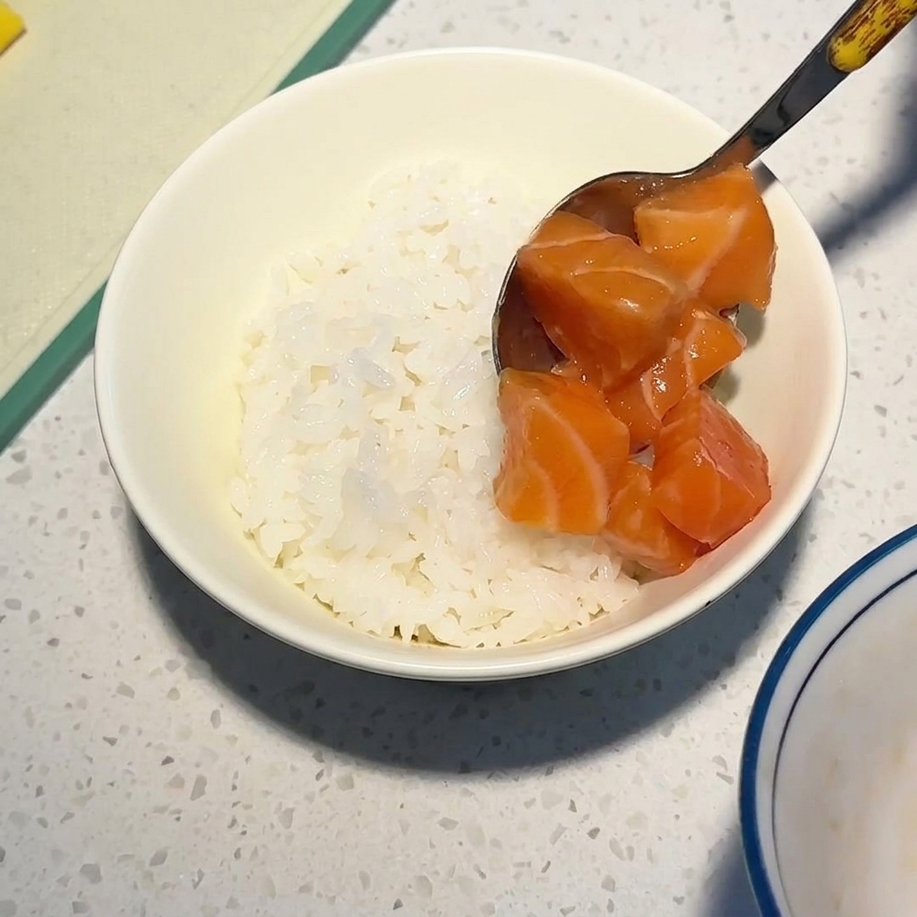 Un cucchiaio di metallo adagia cubetti di salmone crudo lucido su un letto di riso bianco in una ciotola.