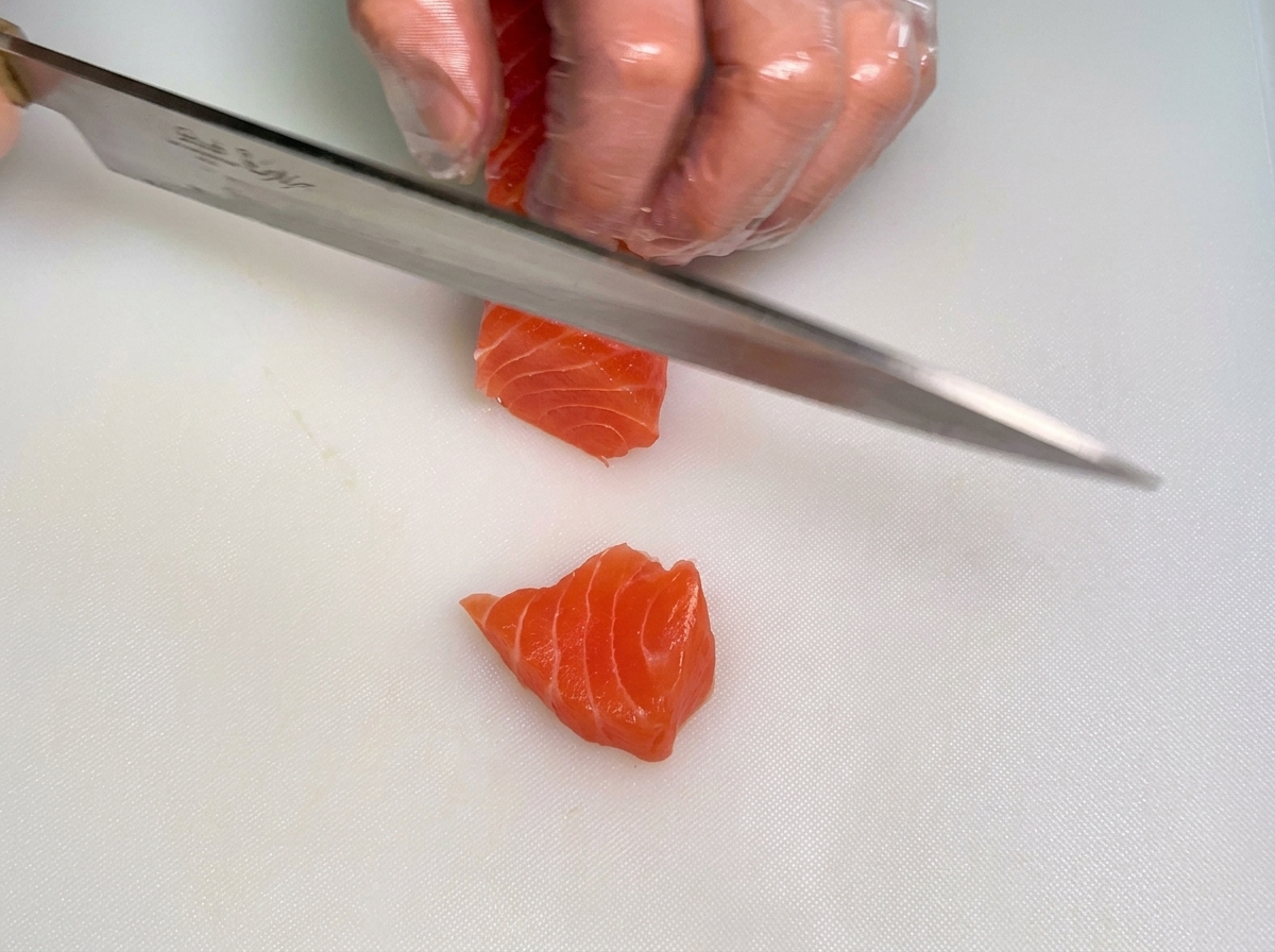 La mano con guanto di uno chef tiene un blocco di salmone crudo mentre laltra mano usa un coltello per tagliarlo a strisce spesse su un tagliere bianco.