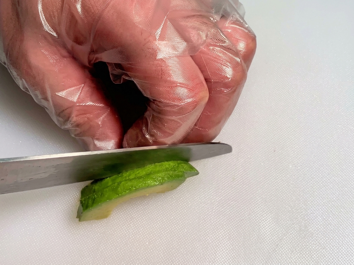Mani con guanti usano un coltello per tagliare a cubetti una fetta di avocado verde fresco.