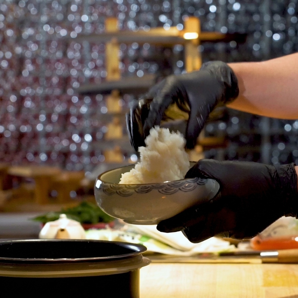 Una persona con guanti neri da cucina che serve riso per sushi soffice in una ciotola di ceramica decorata.