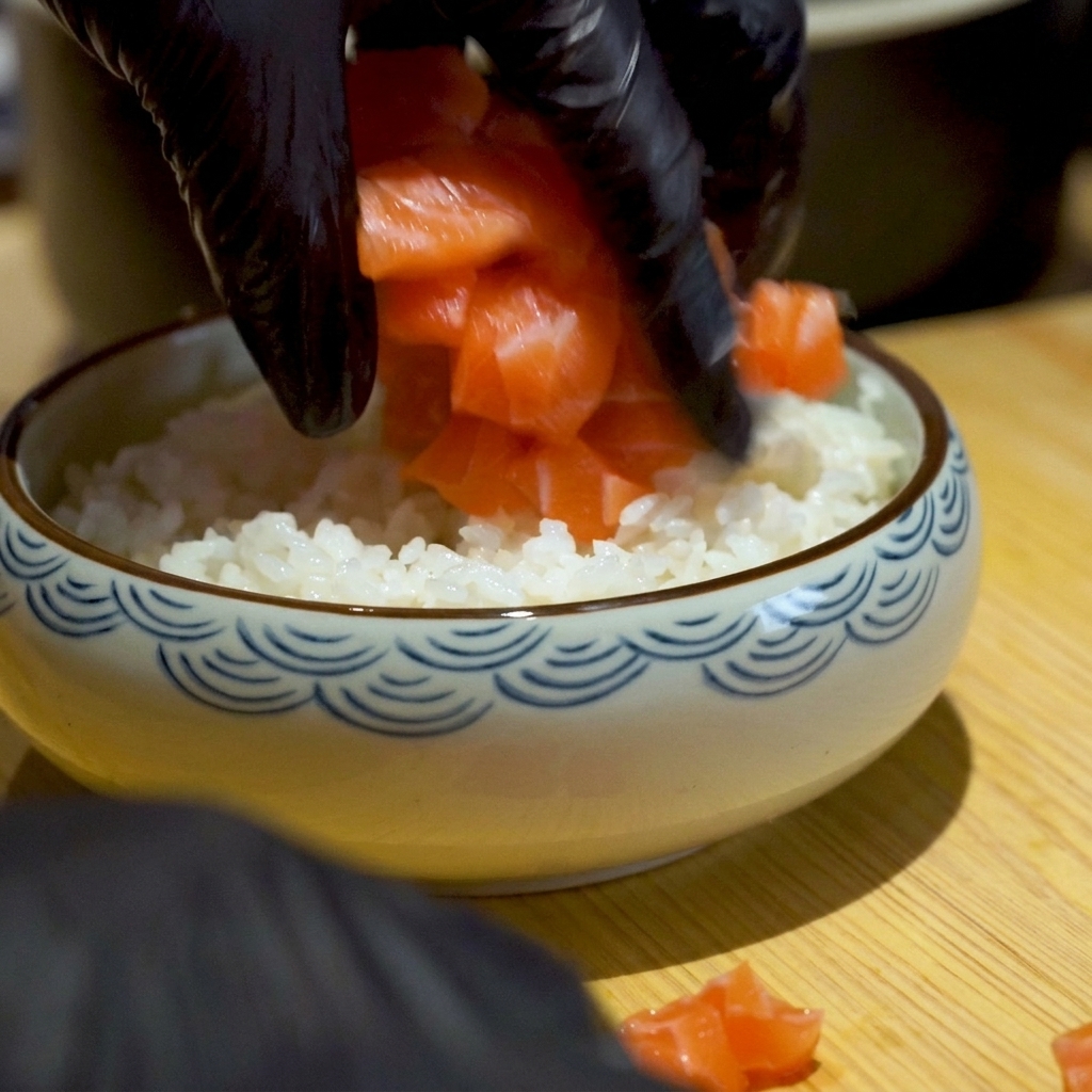 Mani con guanti neri che adagiano cubetti di salmone arancione brillante su un letto di riso bianco in una ciotola.