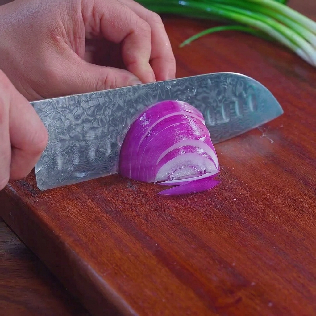 Un coltello da chef che affetta una cipolla rossa viola in strisce sottili su un tagliere di legno, con cipollotti visibili sullo sfondo.