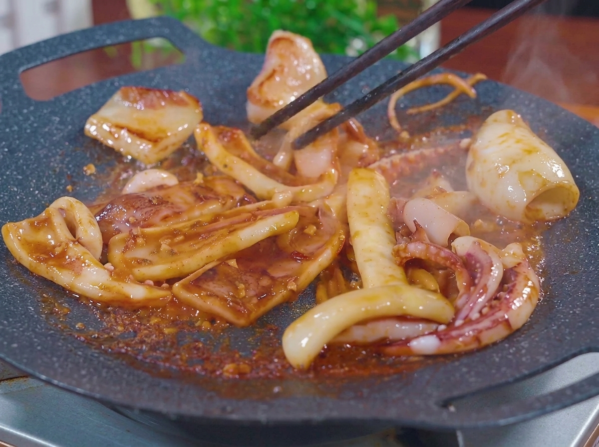 Pezzi di calamaro saltati con le bacchette su una piastra calda e piatta, mentre sfrigolano in una marinatura barbecue densa e spumeggiante marrone-rossastra con vapore che sale.