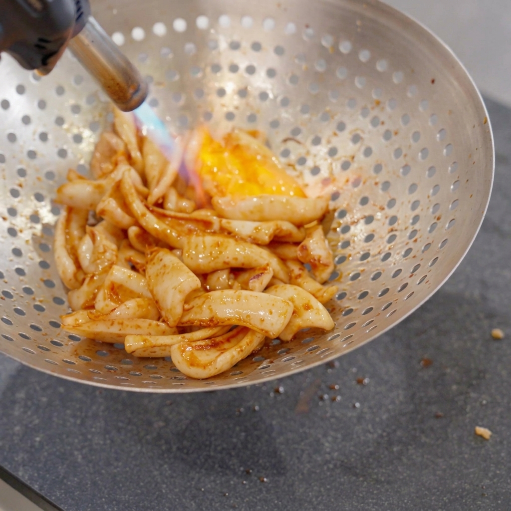 Pezzi di calamaro fritto allinterno di una grande schiumarola metallica forata mentre vengono colpiti da una fiamma blu e arancione di un cannello da cucina.