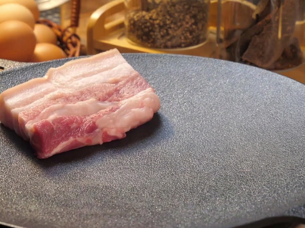 Spesse fette di pancetta di maiale cruda disposte su una piastra nera bollente per essere scottate.