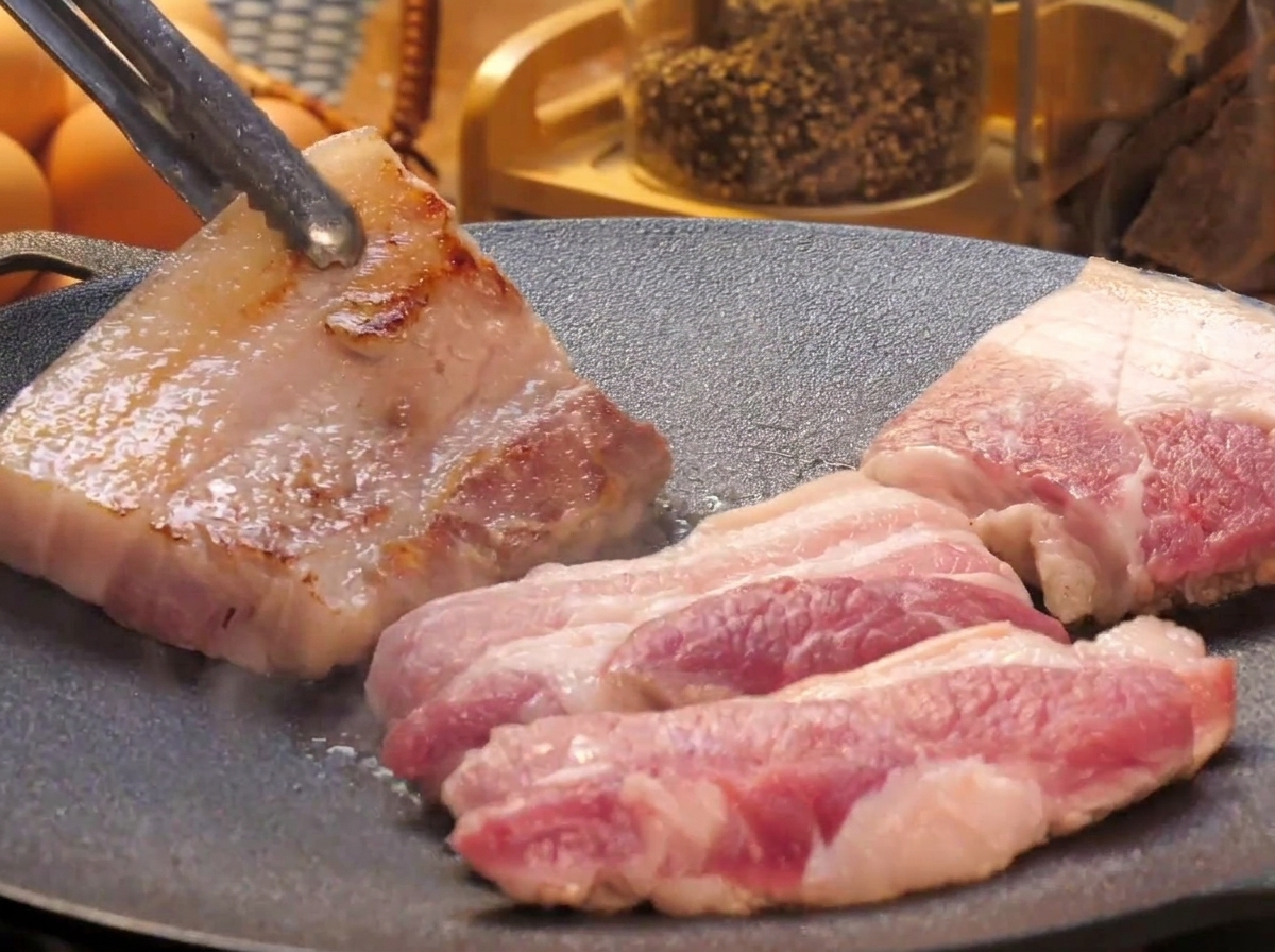 Spesse fette di pancetta che cuociono su una piastra, con un pezzo girato tramite pinze di metallo per mostrare la superficie dorata.