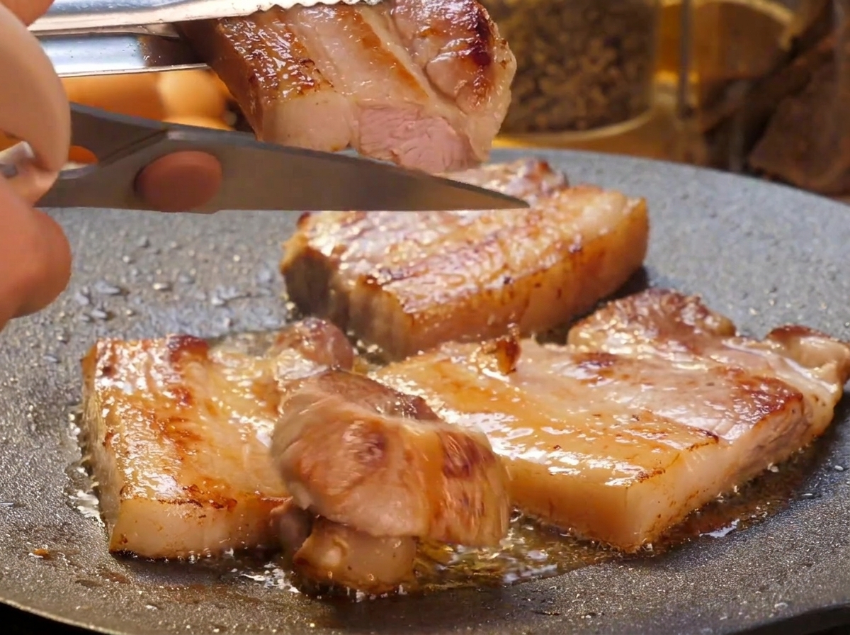 Una mano che usa le forbici da cucina per tagliare le fette di pancetta grigliata in pezzi più piccoli direttamente sulla piastra sfrigolante.
