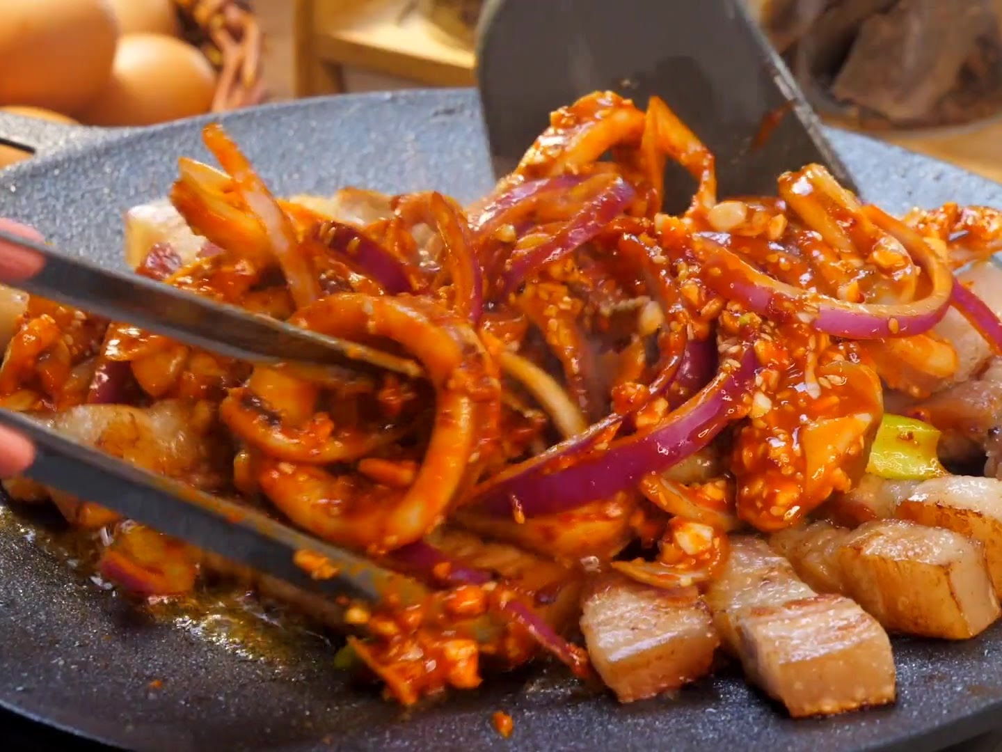 Una spatola e delle pinze che saltano insieme anelli di calamaro marinati, straccetti di pancetta e cipolle rosse su una piastra sfrigolante.