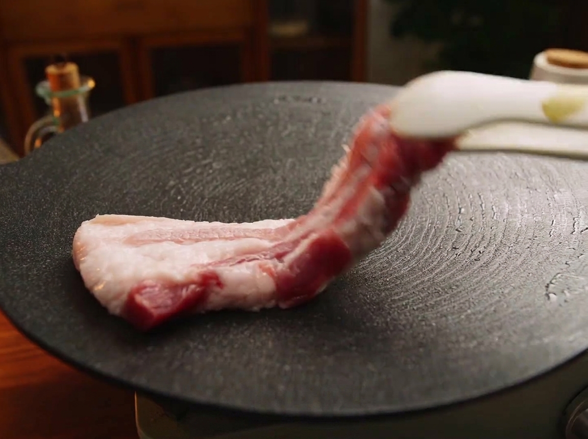 Una pinza da cucina adagia una fetta spessa di pancetta di maiale cruda su una piastra di ghisa nera bollente.