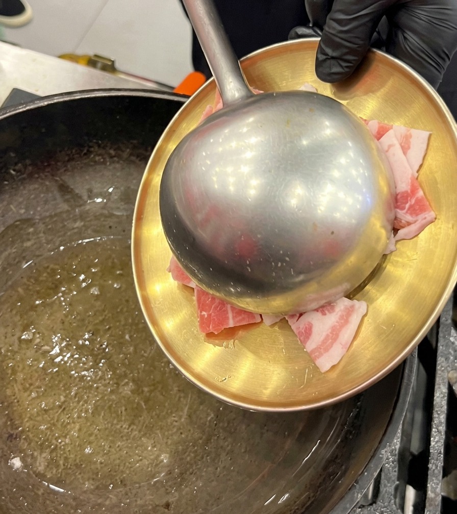 Fette di pancetta cruda che vengono trasferite da un piatto dorato a un wok oliato usando un grande mestolo di metallo.