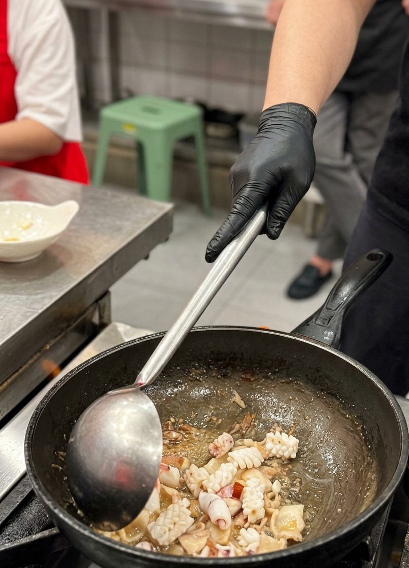 Un primo piano di pezzi di calamaro incisi a incrocio che vengono mescolati in un wok insieme a fette di pancetta rosolate usando un grande mestolo di metallo.