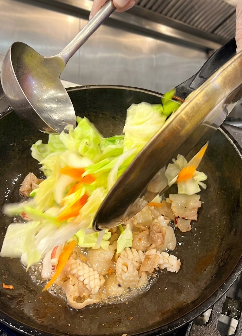 Cavolo cappuccio verde fresco sminuzzato e carote a fette che vengono versati da un piatto di metallo in un wok bollente contenente pancetta e calamari cotti.