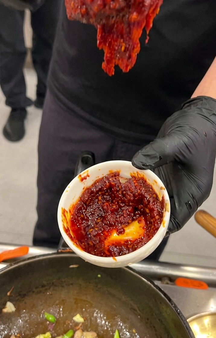 Una mano con un guanto nero che tiene una piccola ciotola bianca piena di una ricca e densa salsa rossa di pasta di peperoncino coreano sopra un wok in cottura.