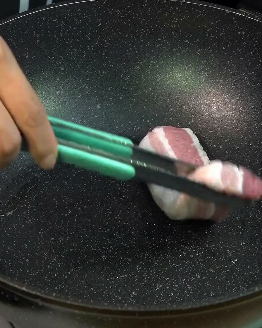 Spesse fette di pancetta di maiale cruda vengono adagiate in una padella nera usando delle pinze verde chiaro per rosolare la carne.
