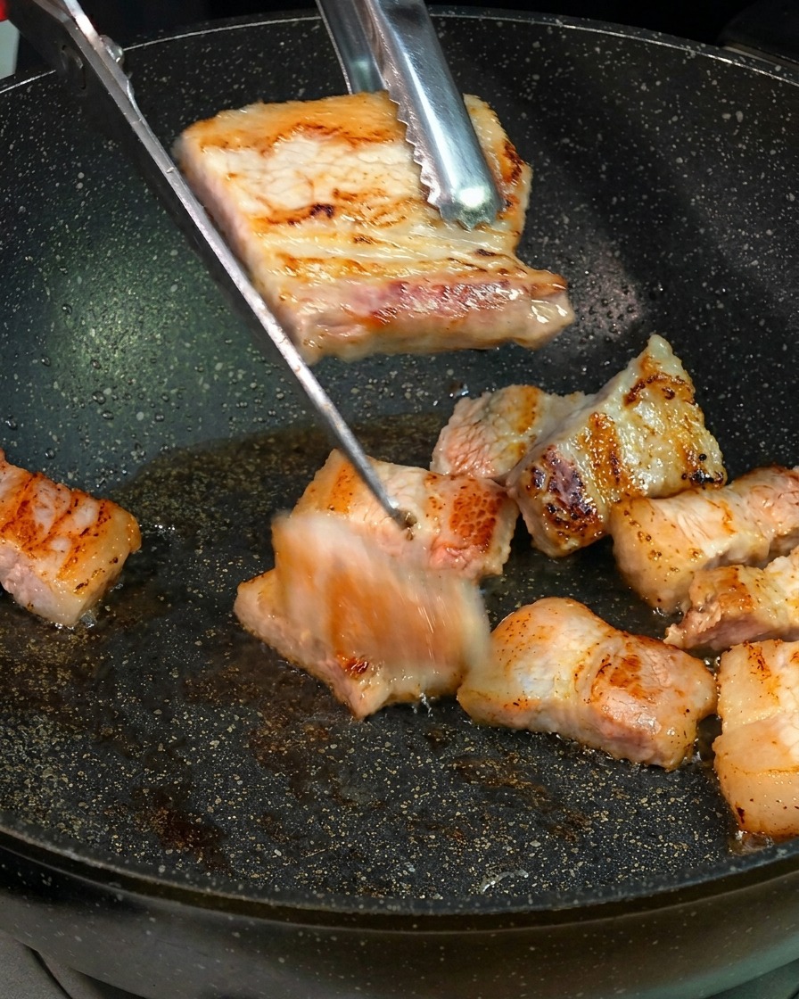 Fette di pancetta di maiale rosolate e dorate vengono tagliate a pezzetti più piccoli con delle forbici da cucina sopra una padella.