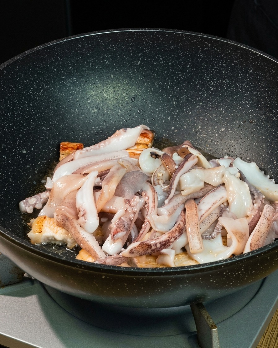 Pezzi di calamaro fresco adagiati sopra i bocconcini di pancetta rosolata allinterno di un profondo wok nero.