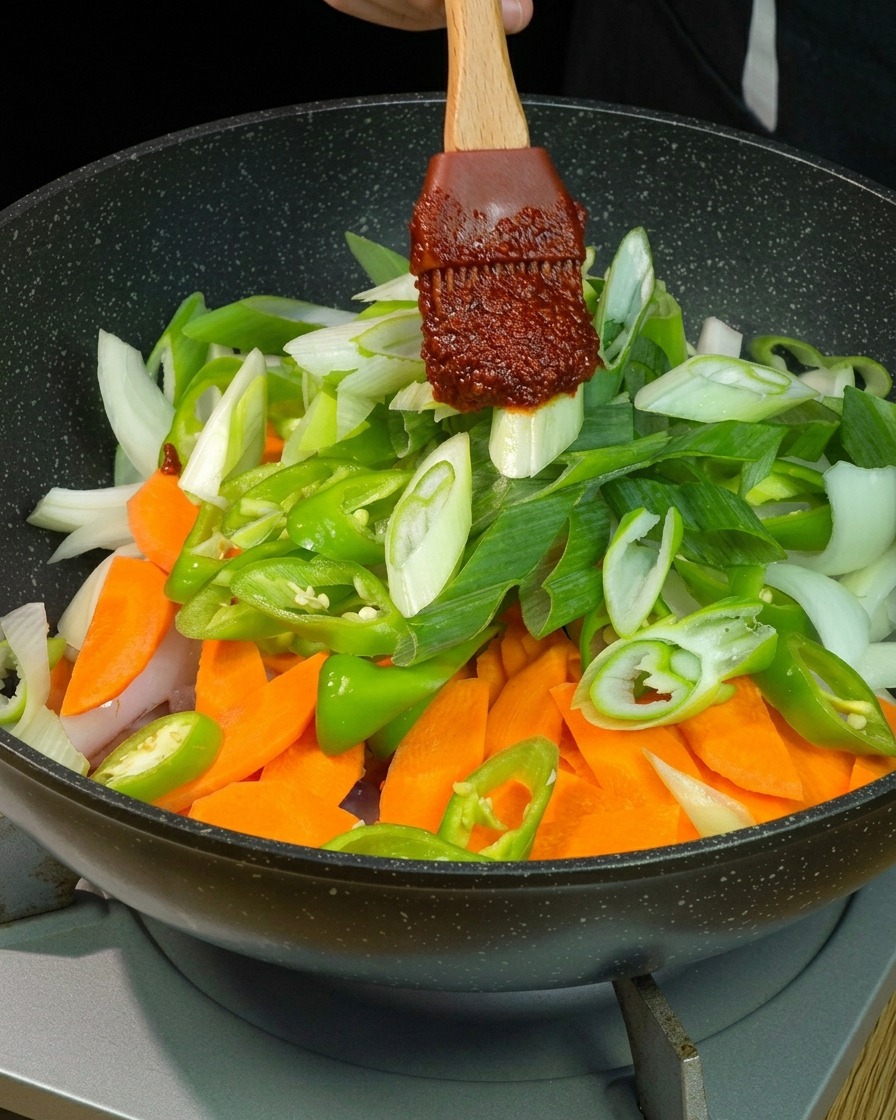 Una generosa cucchiaiata di salsa densa al peperoncino rosso su una spatola di legno viene adagiata sopra una colorata cascata di carote, peperoni verdi, cipolle e cipollotti affettati in padella.