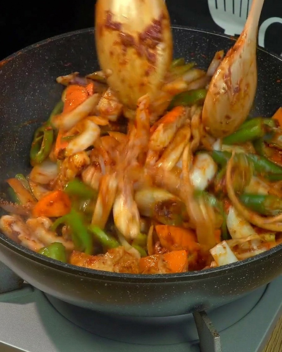 Effetto movimento di pancetta, calamari e verdure miste che vengono fatti saltare energicamente con due cucchiai di legno in un wok, interamente avvolti dalla salsa rossa piccante.