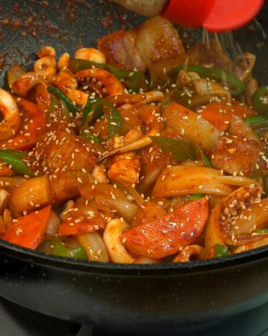 Un primo piano di semi di sesamo bianco che vengono cosparsi da un dosatore con il tappo rosso su un lucido e vibrante spezzatino di calamari, pancetta, carote e peperoni verdi in una padella nera.
