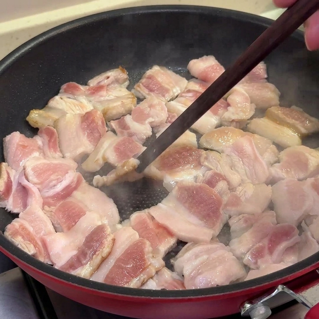 Fette di pancetta di maiale che sfrigolano in una padella antiaderente, diventando dorate e leggermente croccanti, mescolate con le bacchette.