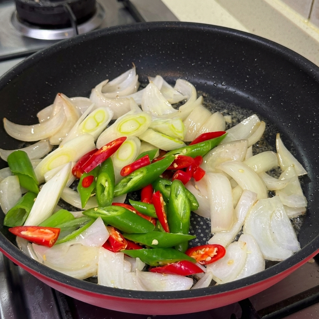 Peperoncini verdi e rossi a fette, cipollotti e aglio tritato che cuociono in padella insieme alle cipolle.