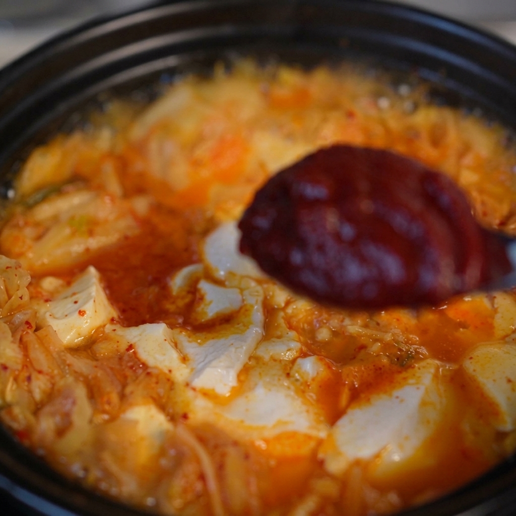 Un cucchiaio con una porzione di densa pasta di peperoncino coreana sopra la zuppa di kimchi e tofu che sobbolle.