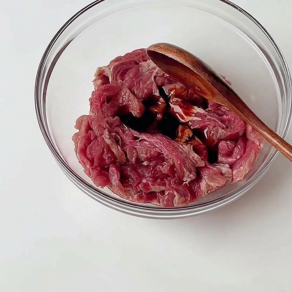 Manzo crudo a fettine sottili in una ciotola di vetro trasparente mescolato con salse scure usando un cucchiaio di legno.