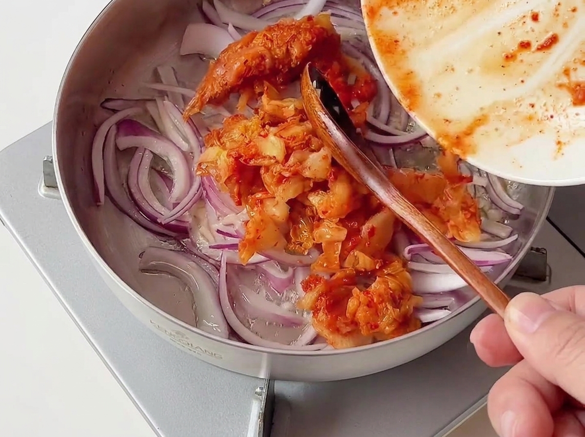 Una porzione di kimchi rosso tritato che viene aggiunta da un piatto in una padella di acciaio inossidabile contenente cipolle rosse affettate.