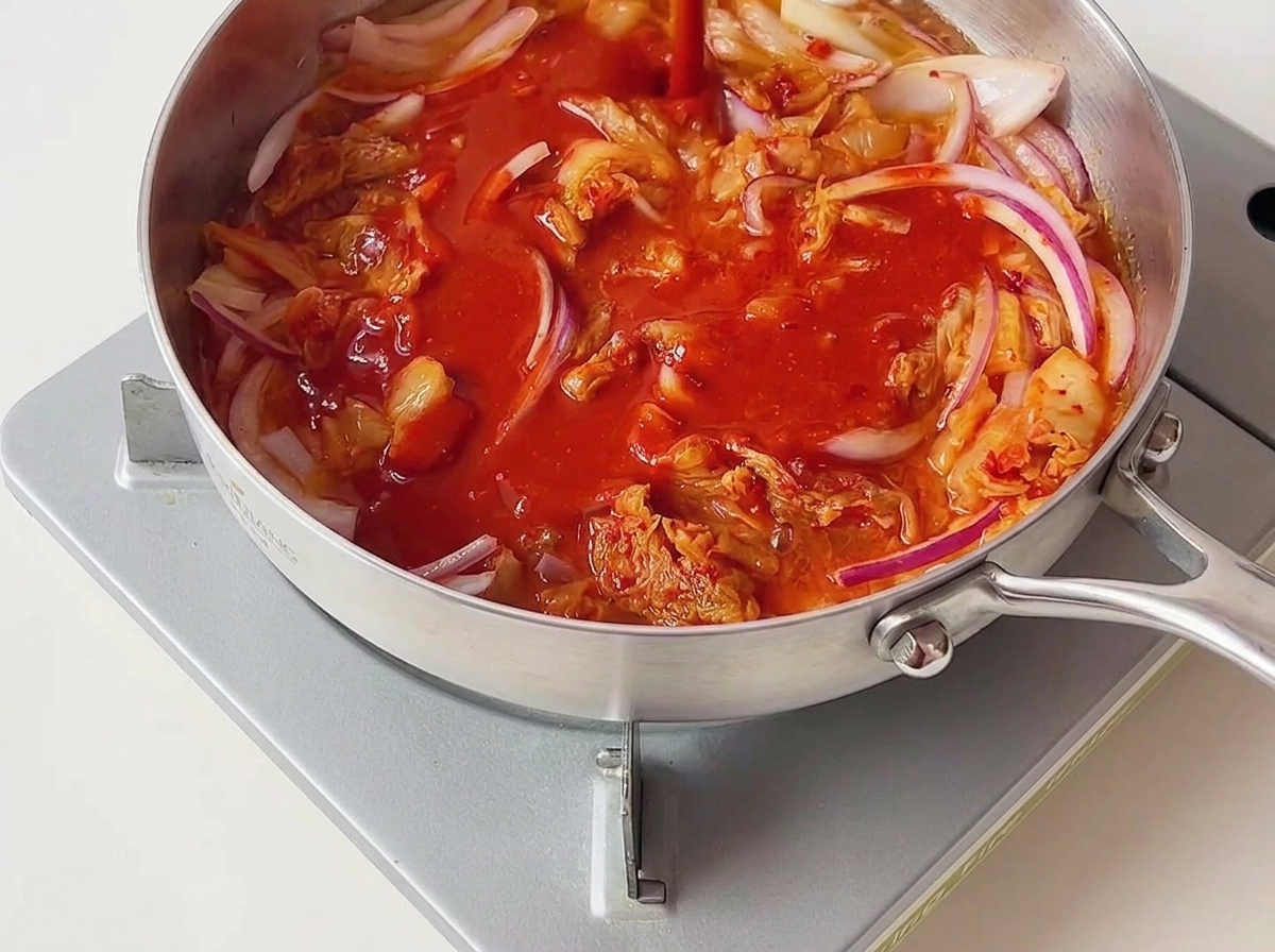 Salsa al peperoncino rosso brillante versata sopra kimchi e cipolle rosse saltati allinterno di una padella dargento.