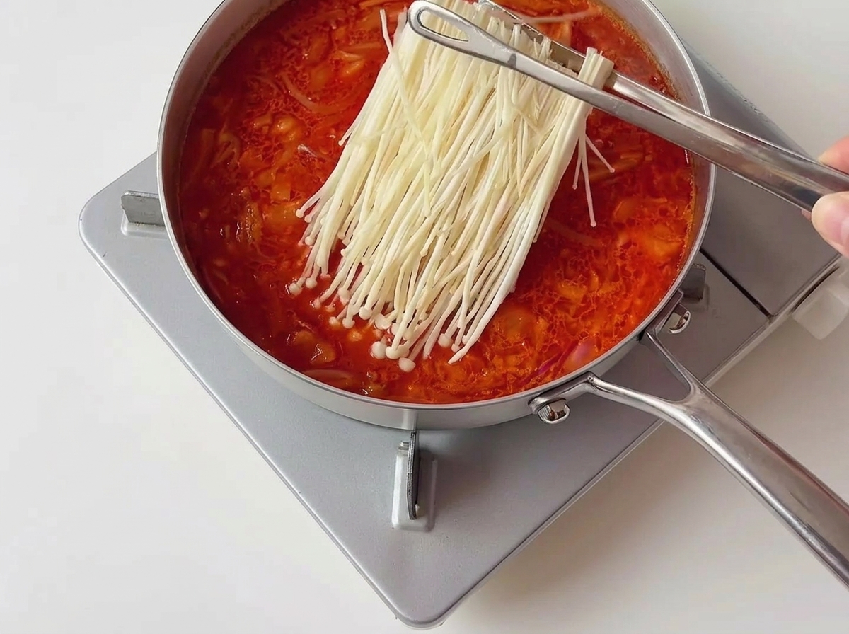Pinze di metallo che posizionano un mazzetto ordinato di funghi enoki bianchi in uno stufato di kimchi rosso bollente.