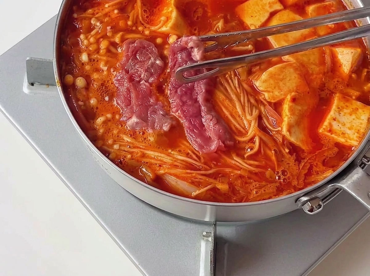Pinze che trasferiscono pezzetti di carne cruda marinata rosa in uno stufato rosso caldo e bollente, accanto a fette di tofu.