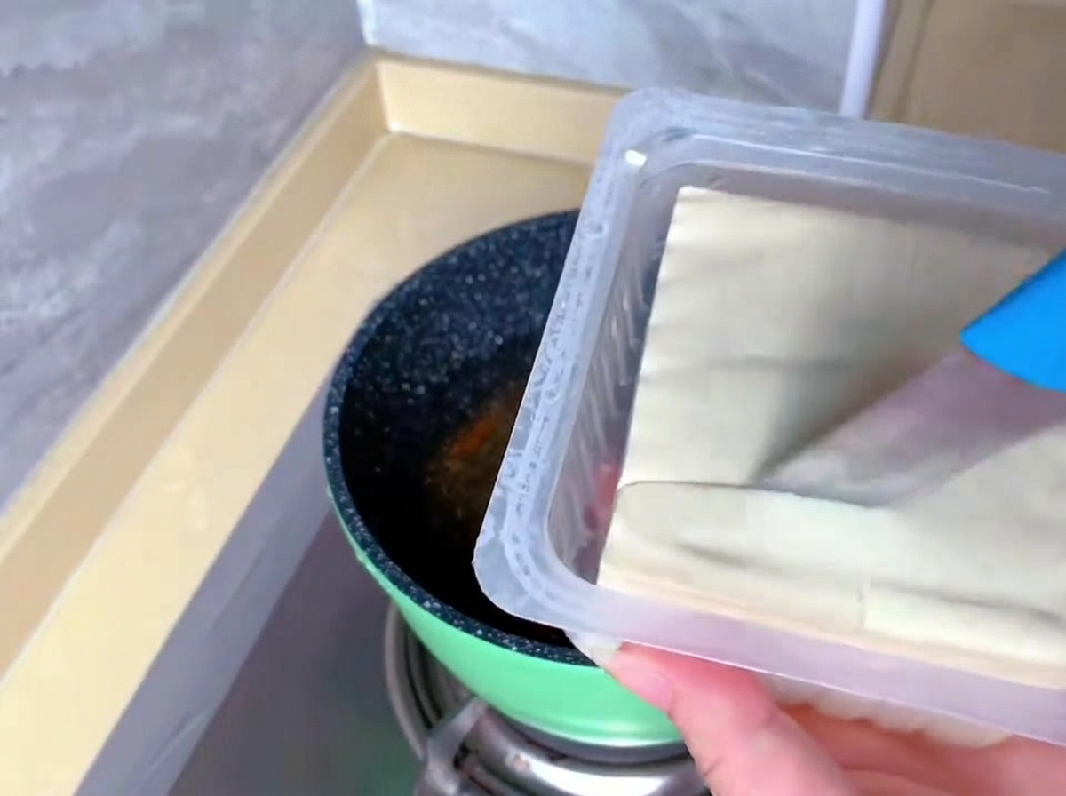 Una persona usa un coltello blu per tagliare un blocco di tofu vellutato bianco direttamente nella sua confezione di plastica.