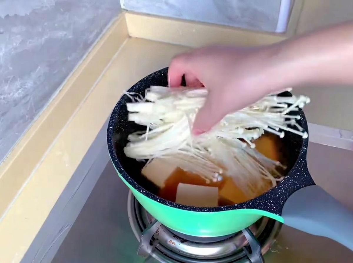 Una mano trasferisce un mazzetto di funghi enoki lunghi e bianchi in una pentola contenente già brodo e tofu.
