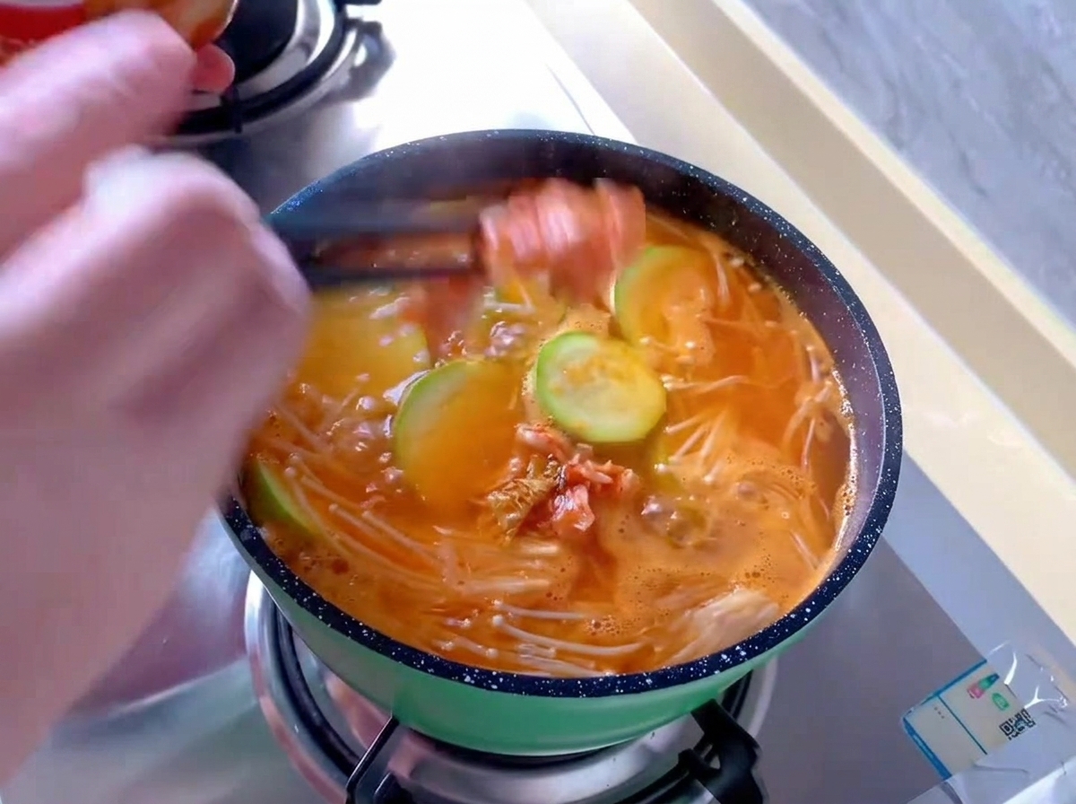 Aggiunta di pezzi di cavolo kimchi rosso con le bacchette in una piccola pentola verde con zuppa arancione che bolle, insieme a funghi enoki e zucchine.