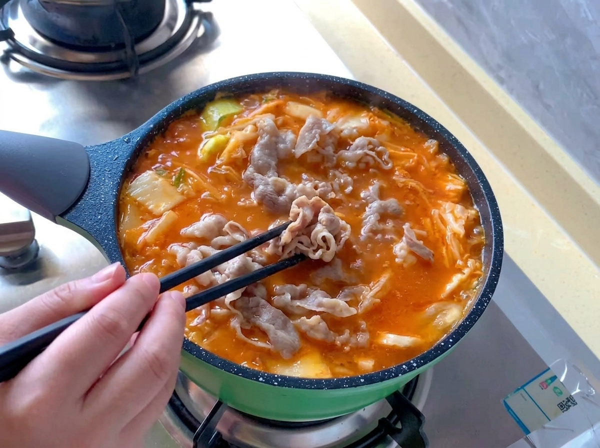 Involtini di manzo sbollentati vengono distribuiti con bacchette nere allinterno di una pentola bollente di zuppa di kimchi rosso-arancio.