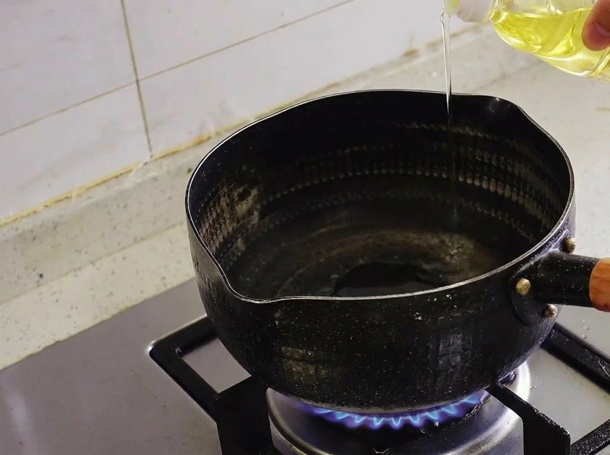 Olio limpido versato da una bottiglia in una pentola nera su un fornello a gas acceso.