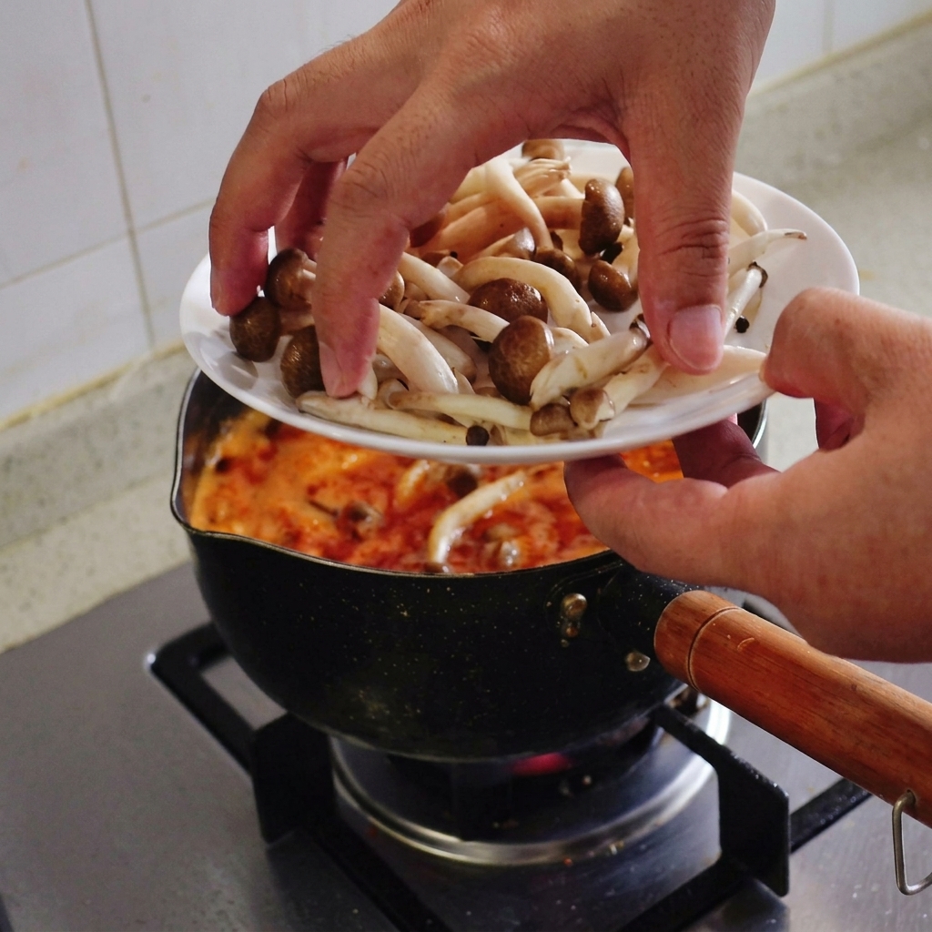 Mani che trasferiscono una manciata di funghi shimeji freschi con il cappello marrone da un piatto bianco a una pentola di zuppa rossa in ebollizione.