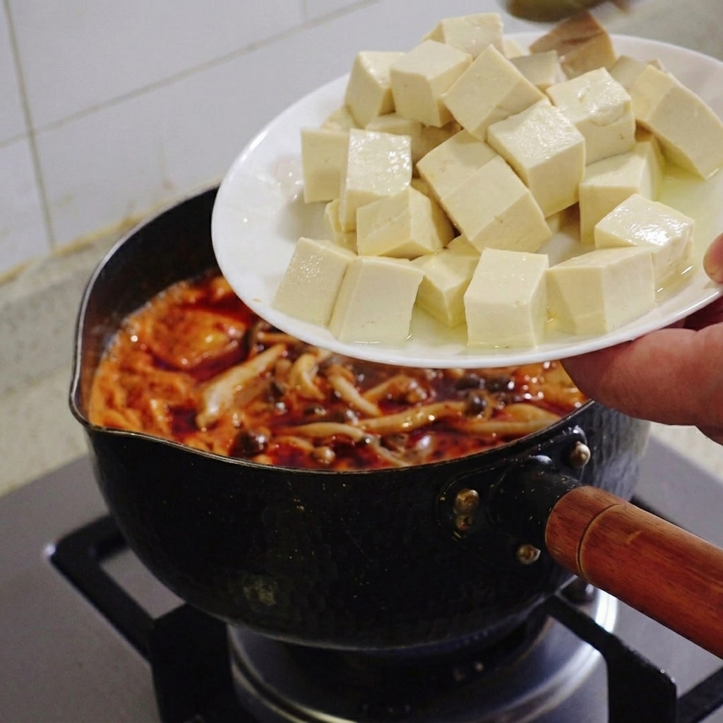 Una mano che tiene un piatto bianco e fa scivolare cubetti regolari di tofu bianco morbido in una pentola di stufato di kimchi rosso che sobbolle.