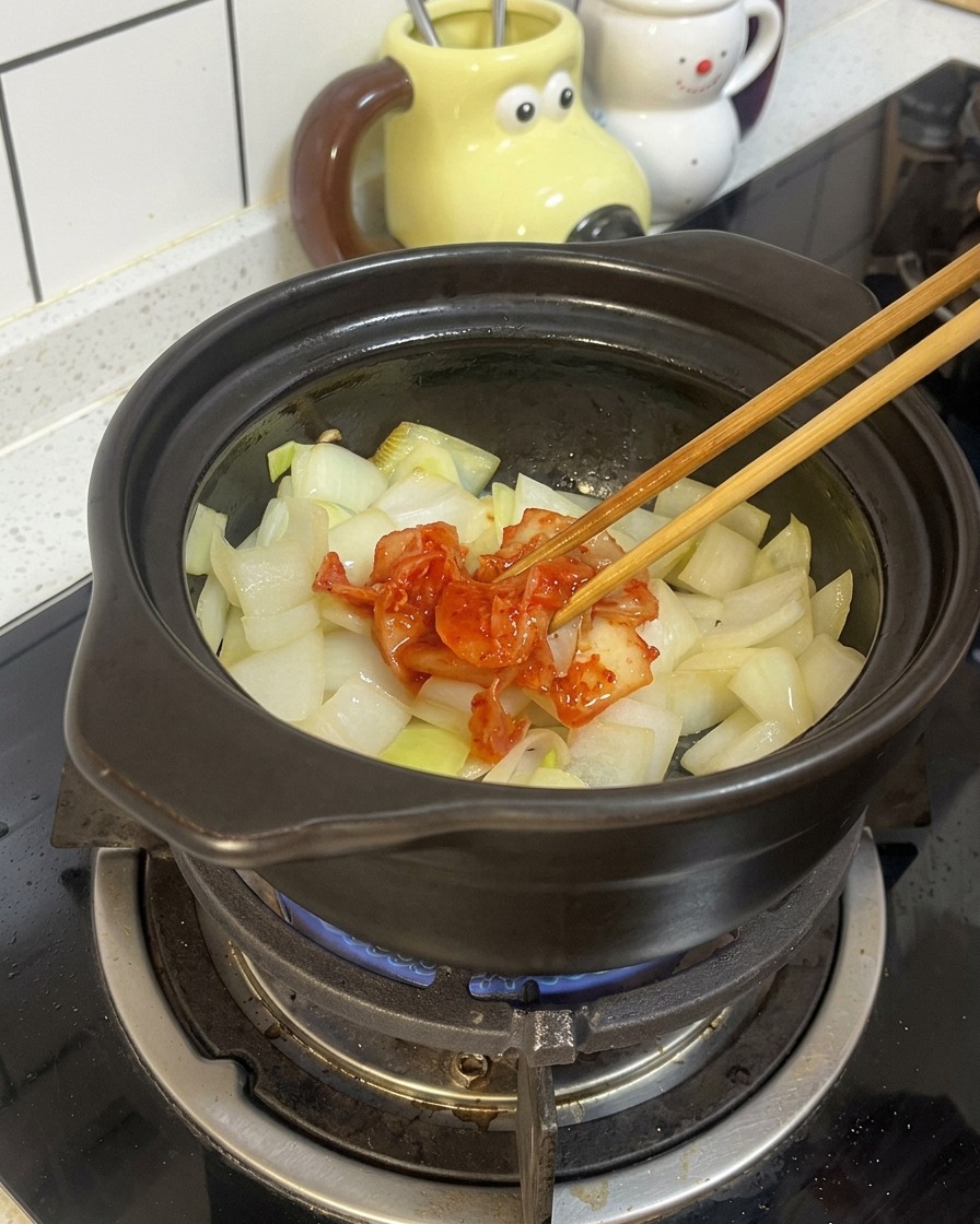 Bacchette di legno posizionano una porzione di kimchi rosso e fermentato sopra le cipolle soffritte in una pentola di terracotta nera.