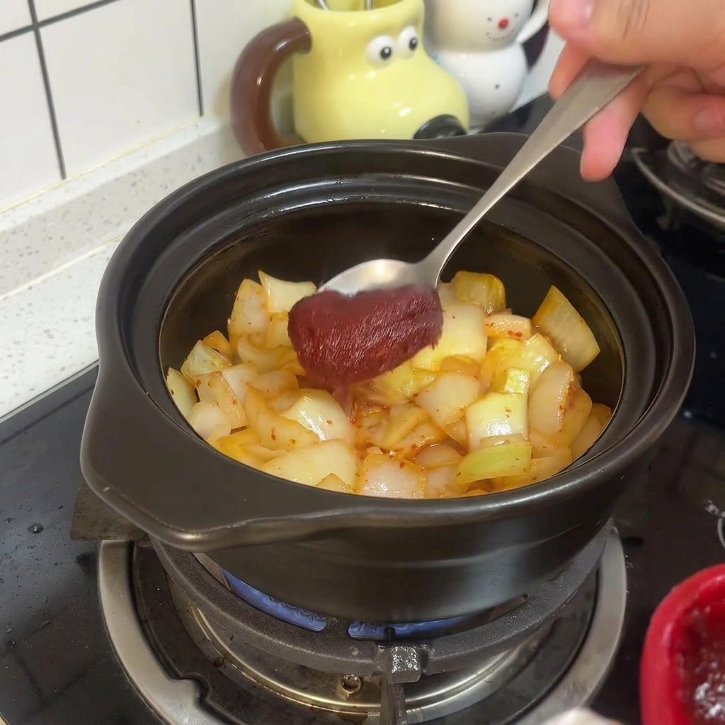 Un cucchiaio di metallo mescola la densa pasta di peperoncino rosso coreano al kimchi e alle cipolle soffritte nella pentola.