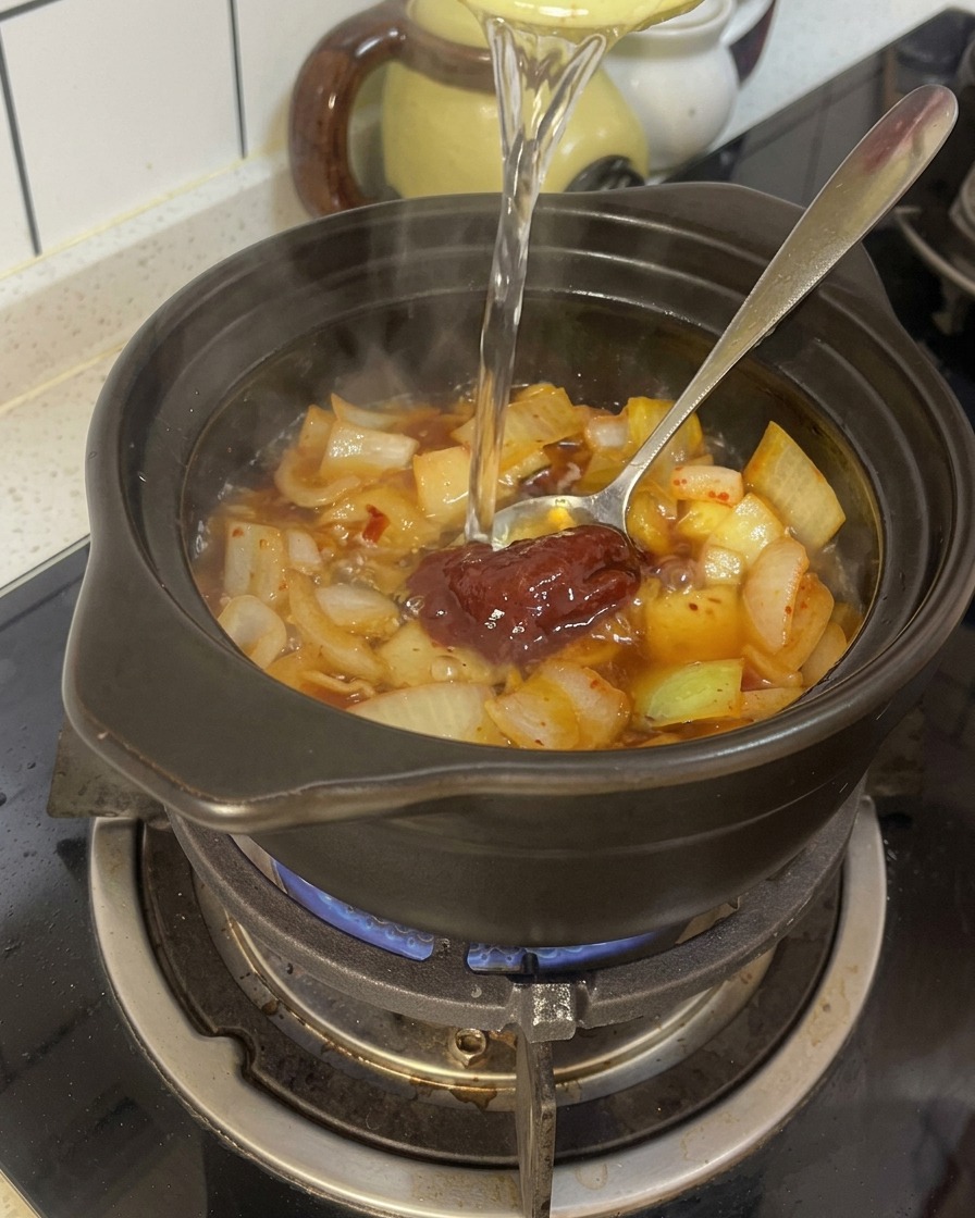 Acqua versata in una pentola di terracotta nera che contiene cipolle condite e pasta di peperoncino rosso.