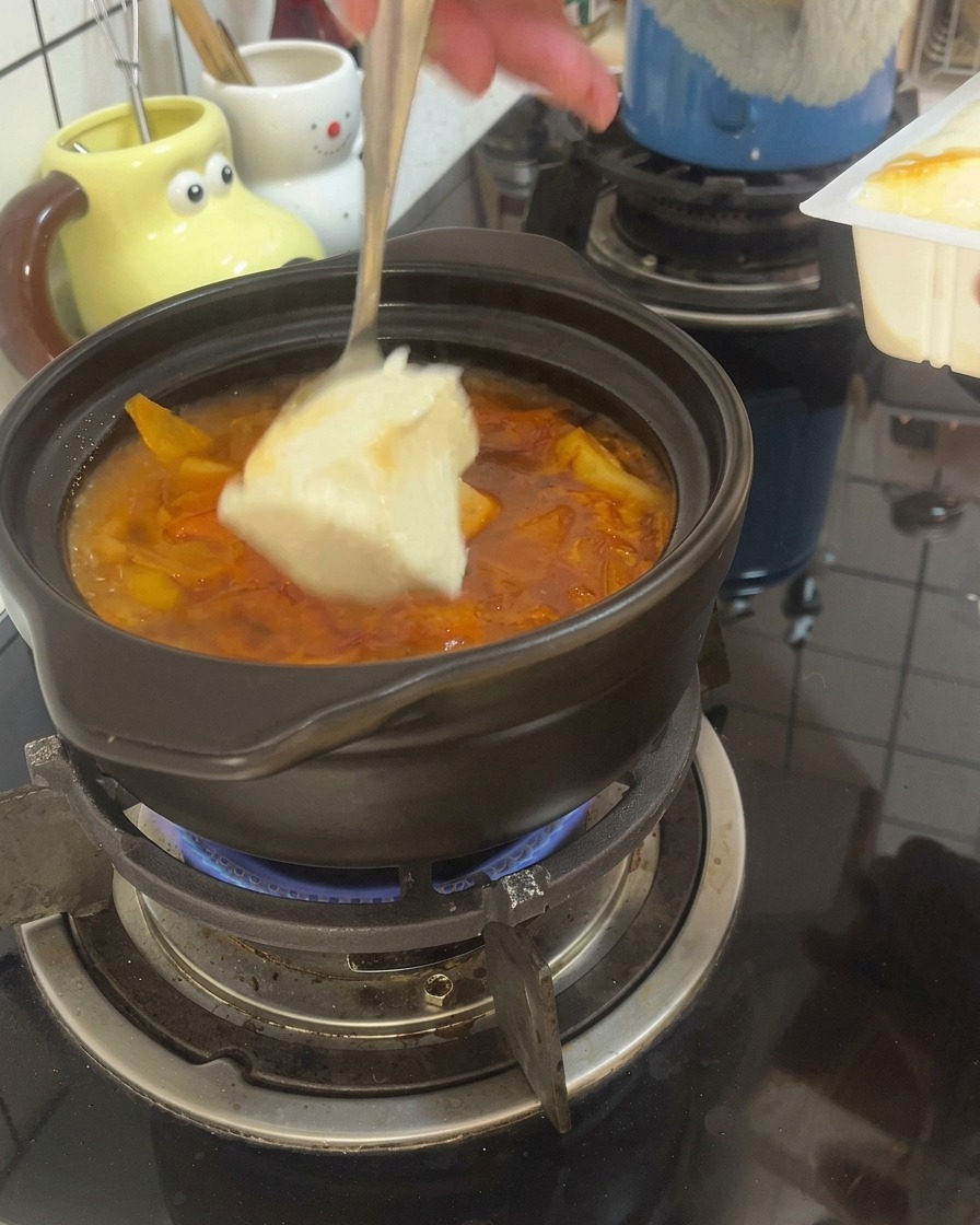Un cucchiaio versa grossi pezzi di tofu morbido nel brodo rosso che ribolle in una pentola di terracotta.