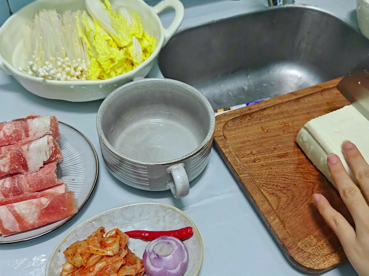 Un primo piano di una mano che usa un coltello da cucina per tagliare un grande blocco di tofu bianco in spessi pezzi rettangolari su un tagliere di legno.