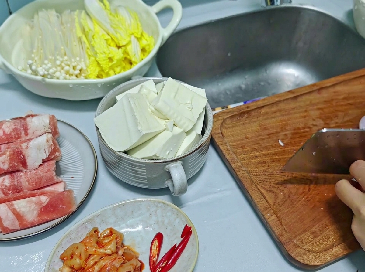 Una mannaia da cucina utilizzata per tritare la cipolla rossa e il peperoncino su un tagliere di legno, con il tofu a fette visibile in una ciotola di lato.