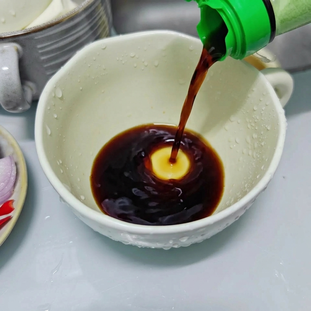Salsa di soia chiara versata da una bottiglia con tappo verde in una tazza di ceramica bianca per preparare il condimento.