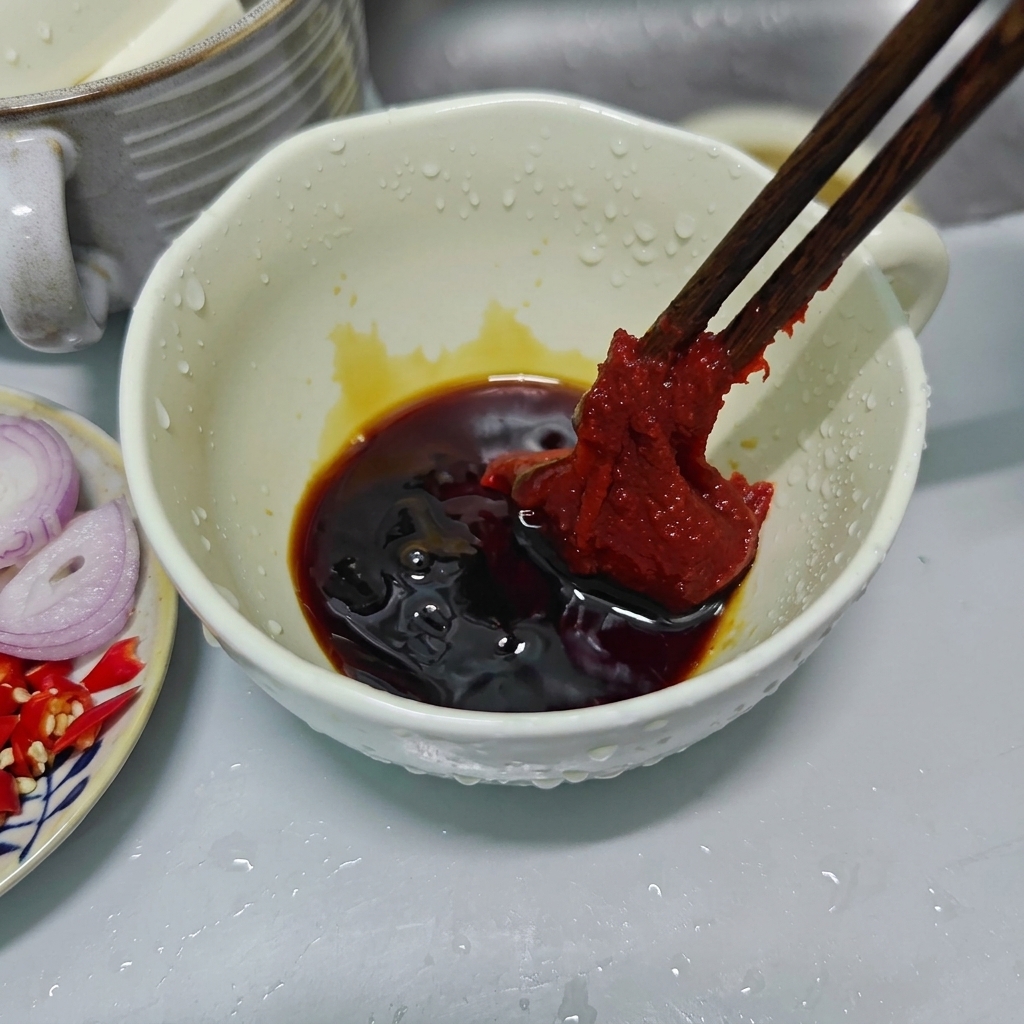 Bacchette di legno che tengono una generosa cucchiaiata di densa pasta di peperoncino coreano rosso sopra una ciotola di ceramica bianca con salse scure.