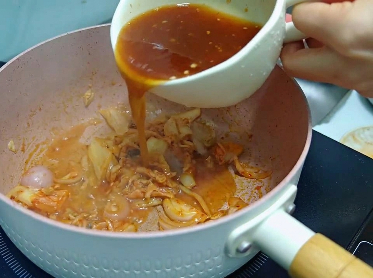 Una persona che versa una salsa mista rossastra da una tazza bianca in una padella rosa contenente kimchi e cipolle saltate.