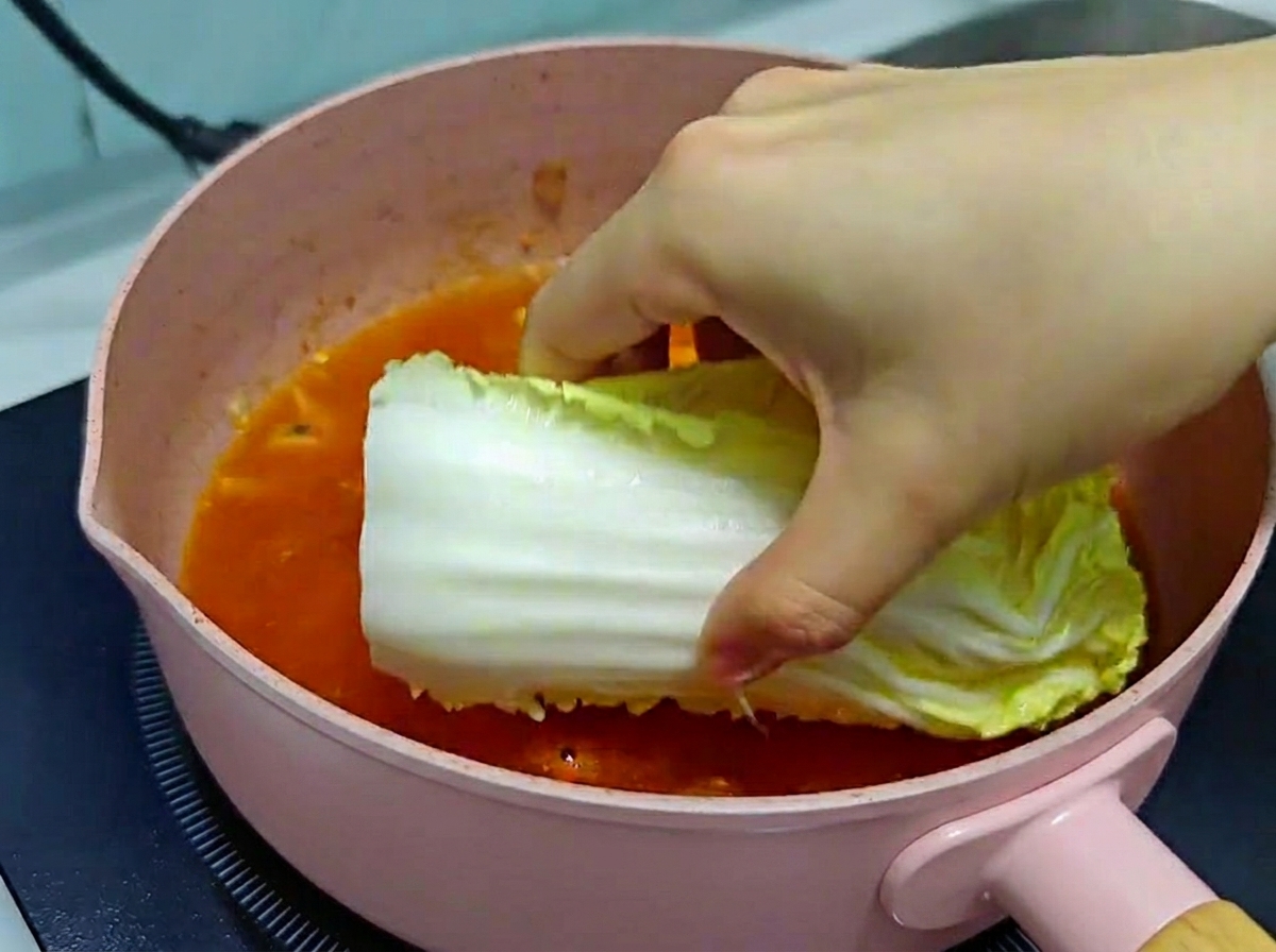 Una mano che adagia con cura un pezzo di cavolo cinese fresco in una padella rosa piena di brodo di zuppa di kimchi che bolle.